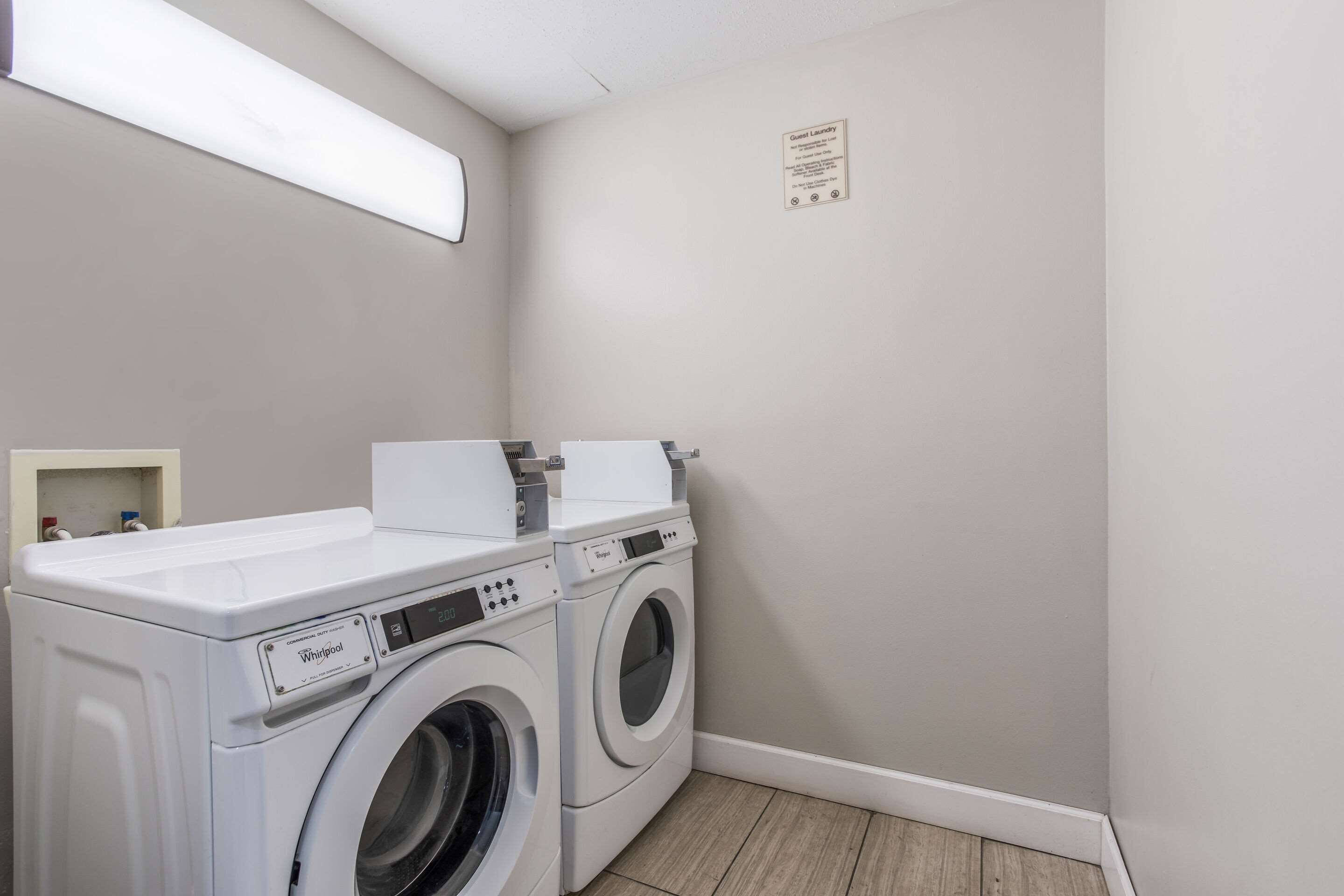 Une buanderie équipée de deux machines à laver et deux sèche-linge Whirlpool, d’une petite étagère contenant des articles de toilette et d’un panneau affiché au mur.