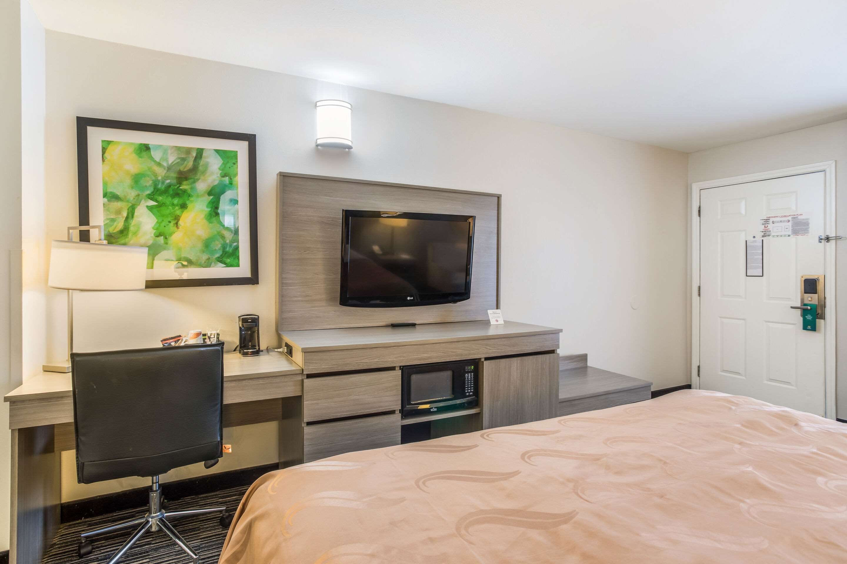 A hotel room with a bed, a desk with a chair, a flat-screen TV, a microwave, and a door with a lock.