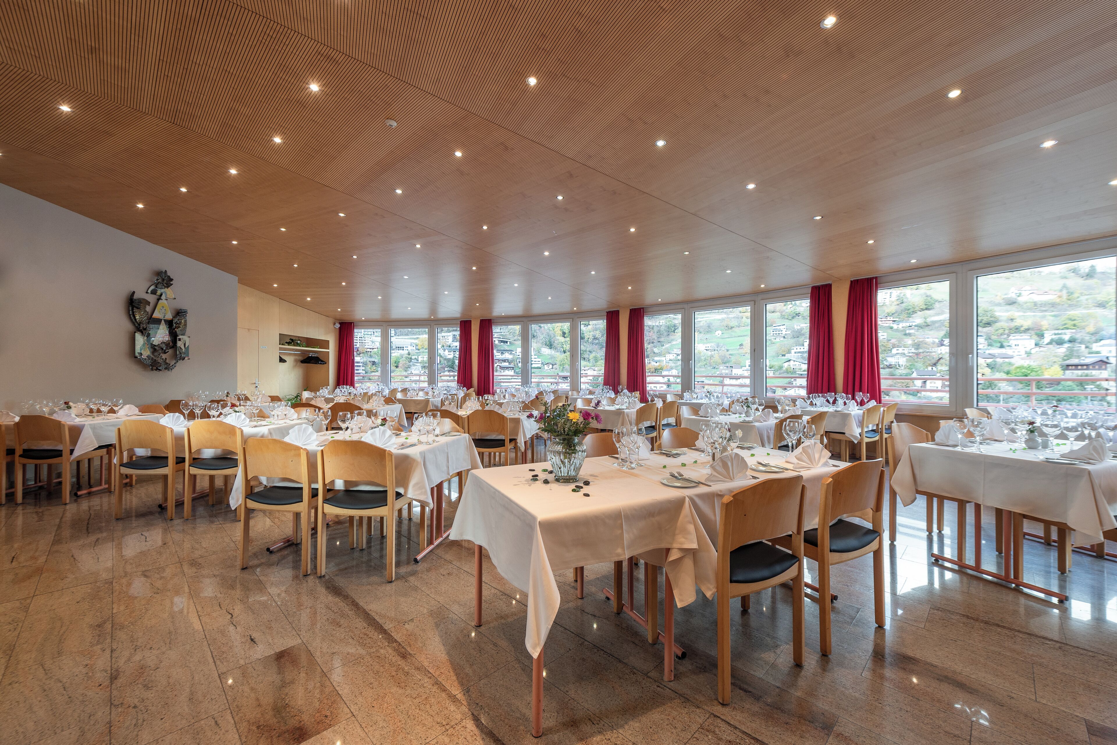 A spacious dining area with multiple tables set for a meal, large windows with red curtains, and a wooden ceiling.