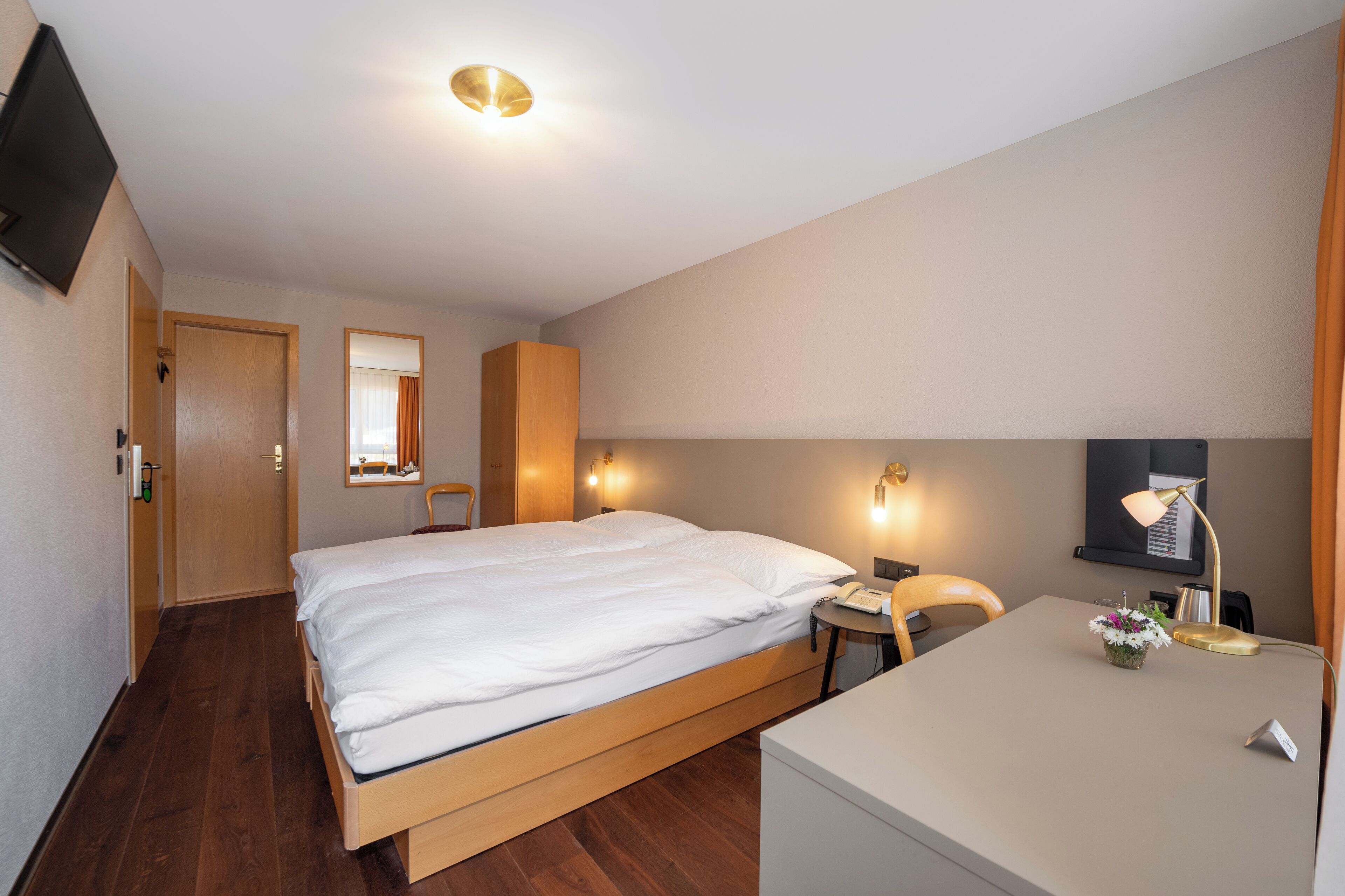 A hotel room with a bed, bedside table, lamp, and a small desk with a chair.