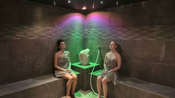 Um espaço de spa com duas pessoas sentadas em bancos, uma decoração rochosa central e iluminação verde ambiente.