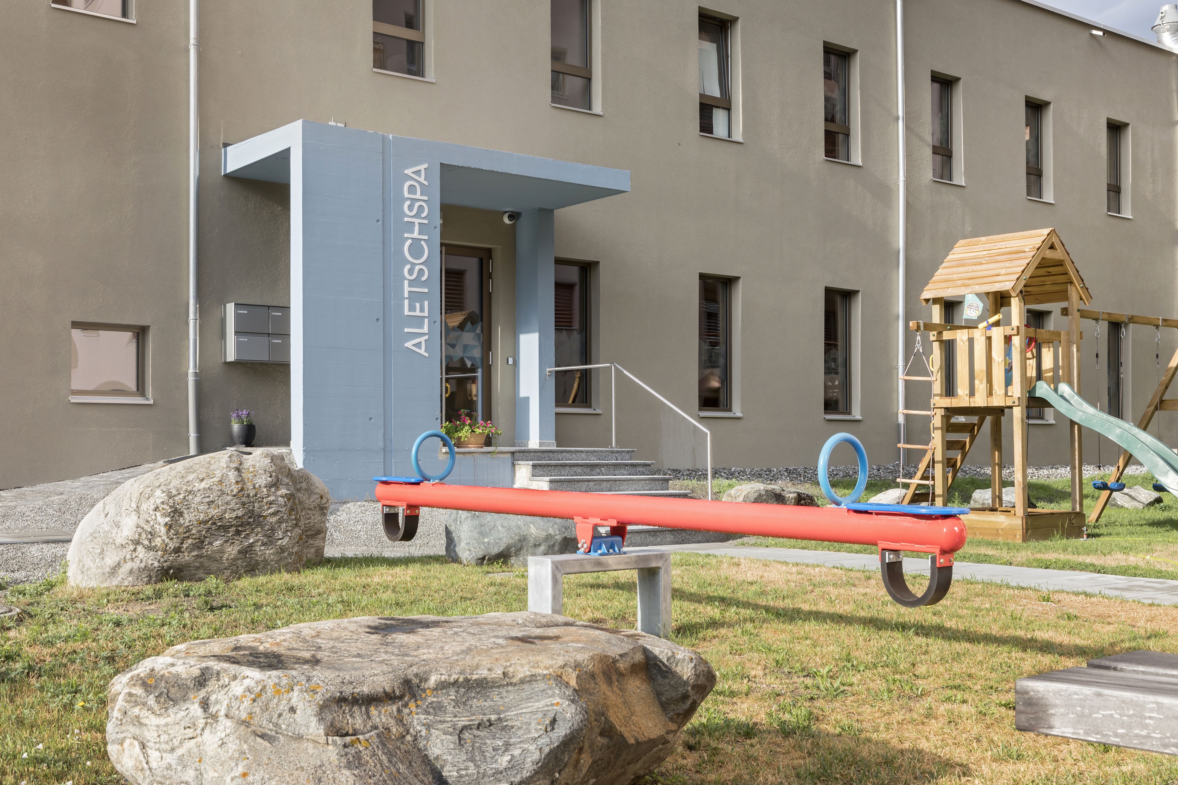 A playground with a seesaw, a wooden playhouse, and a building labeled "ALETSCHSPA" in the background.