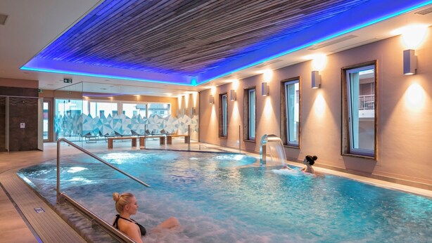 A large indoor swimming pool with a wooden ceiling and blue lighting, surrounded by seating areas and windows. - Hotel Alex (Naters)