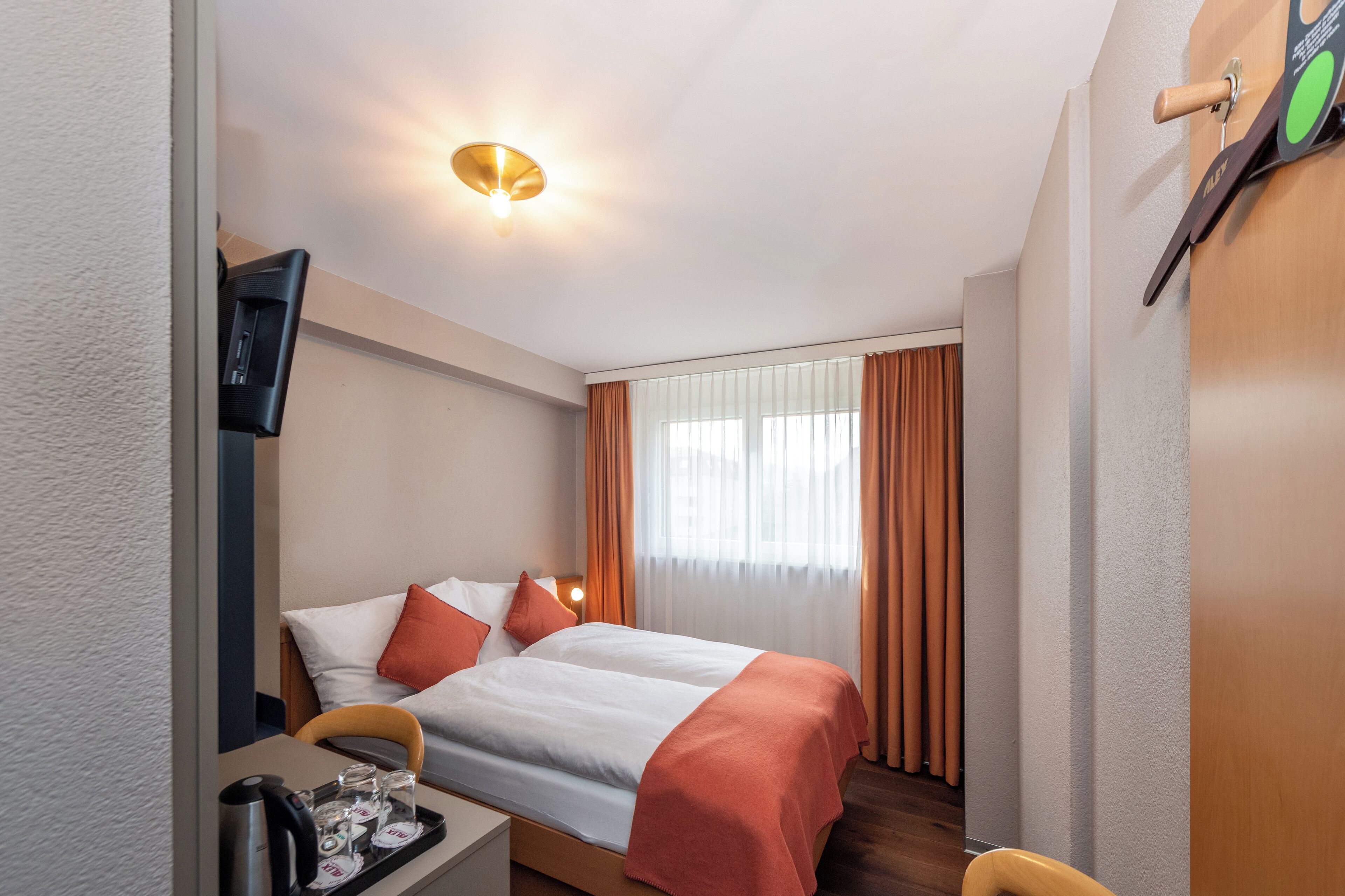 A hotel room with a bed, a desk with a tray of items, a television, and a window with curtains.