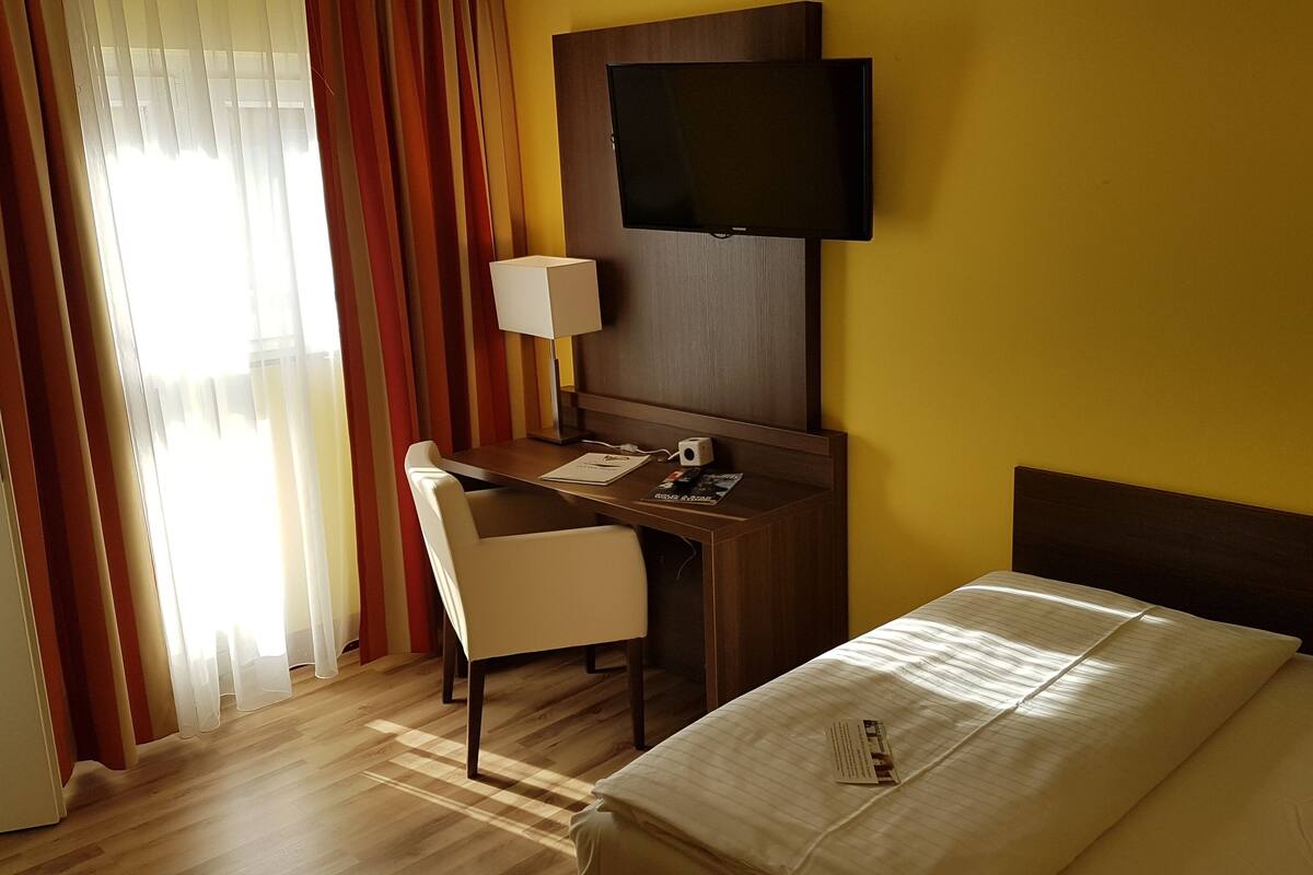 A hotel room with a bed, a desk with a chair, a television, and a window with curtains.