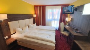 Quarto de hotel com duas camas, uma mesa, uma cadeira, uma televisão e uma janela com cortinas.