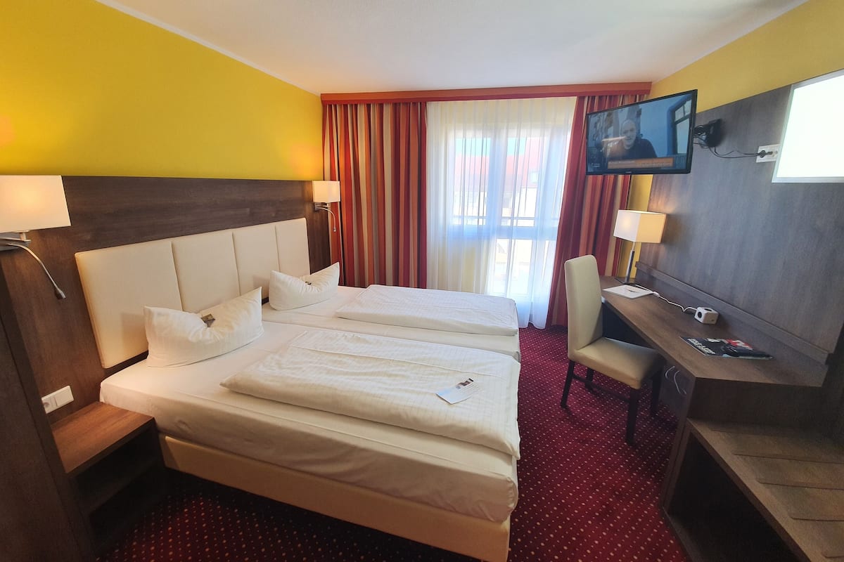 A hotel room with two beds, a desk, a chair, a TV, and a window with curtains.