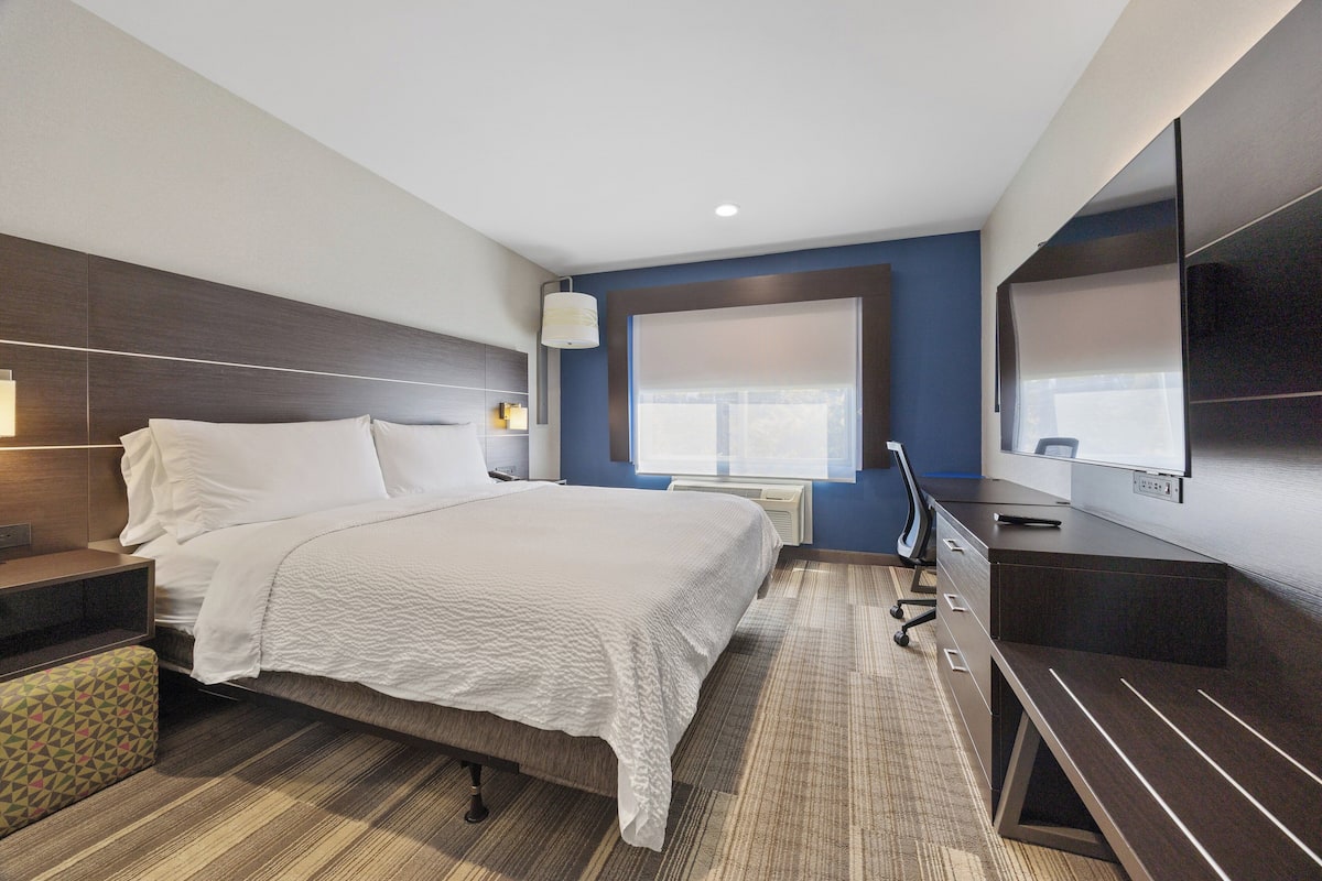 Quarto de hotel com cama, escrivaninha, televisão e janela com persianas.