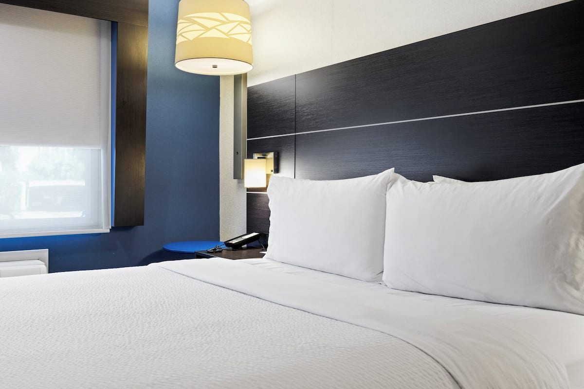 Quarto de hotel com uma cama grande, uma mesa de cabeceira, uma luminária e uma janela com persianas.