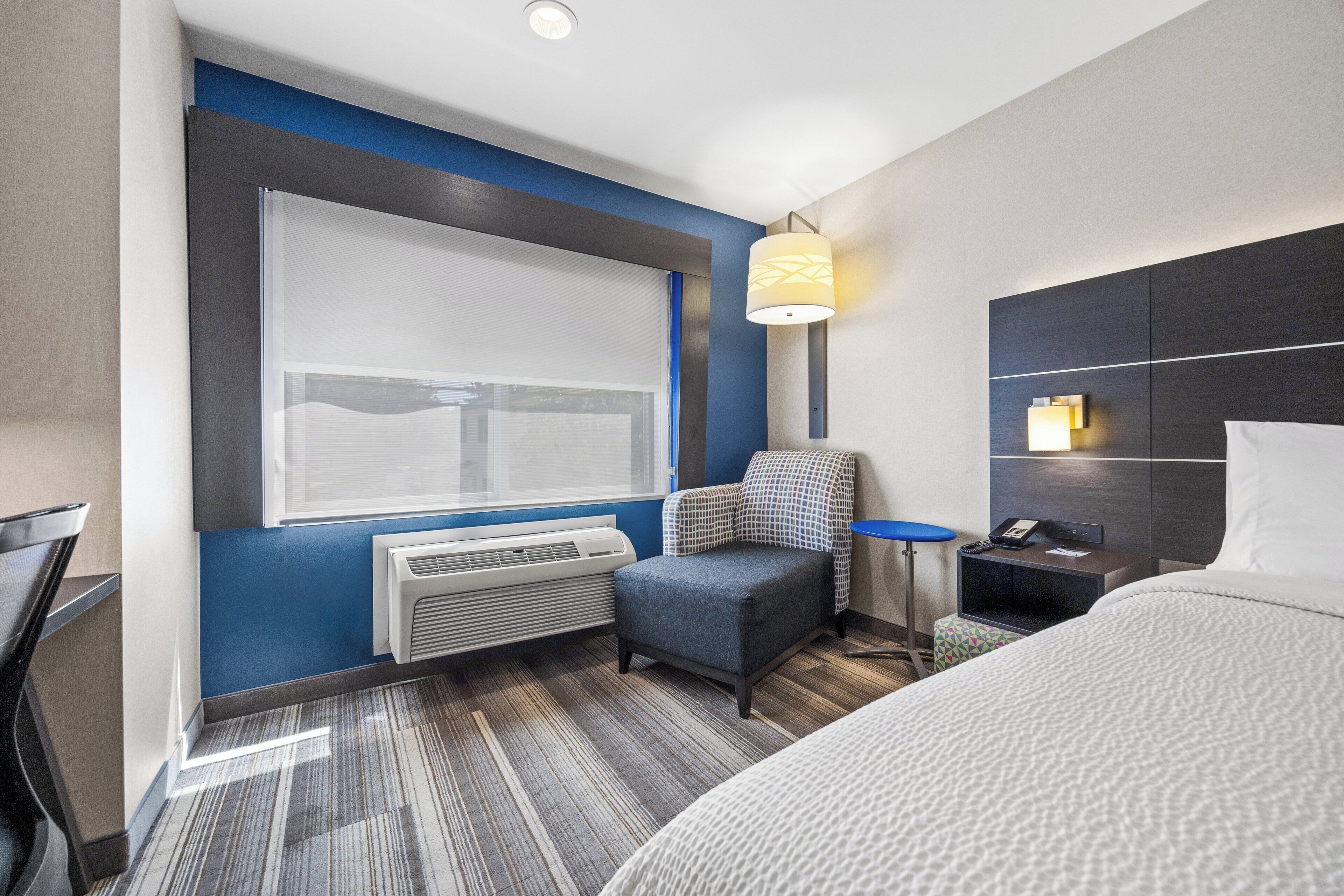 A modern hotel room with a large bed, a desk, a chair, a small table, and a wall-mounted lamp.