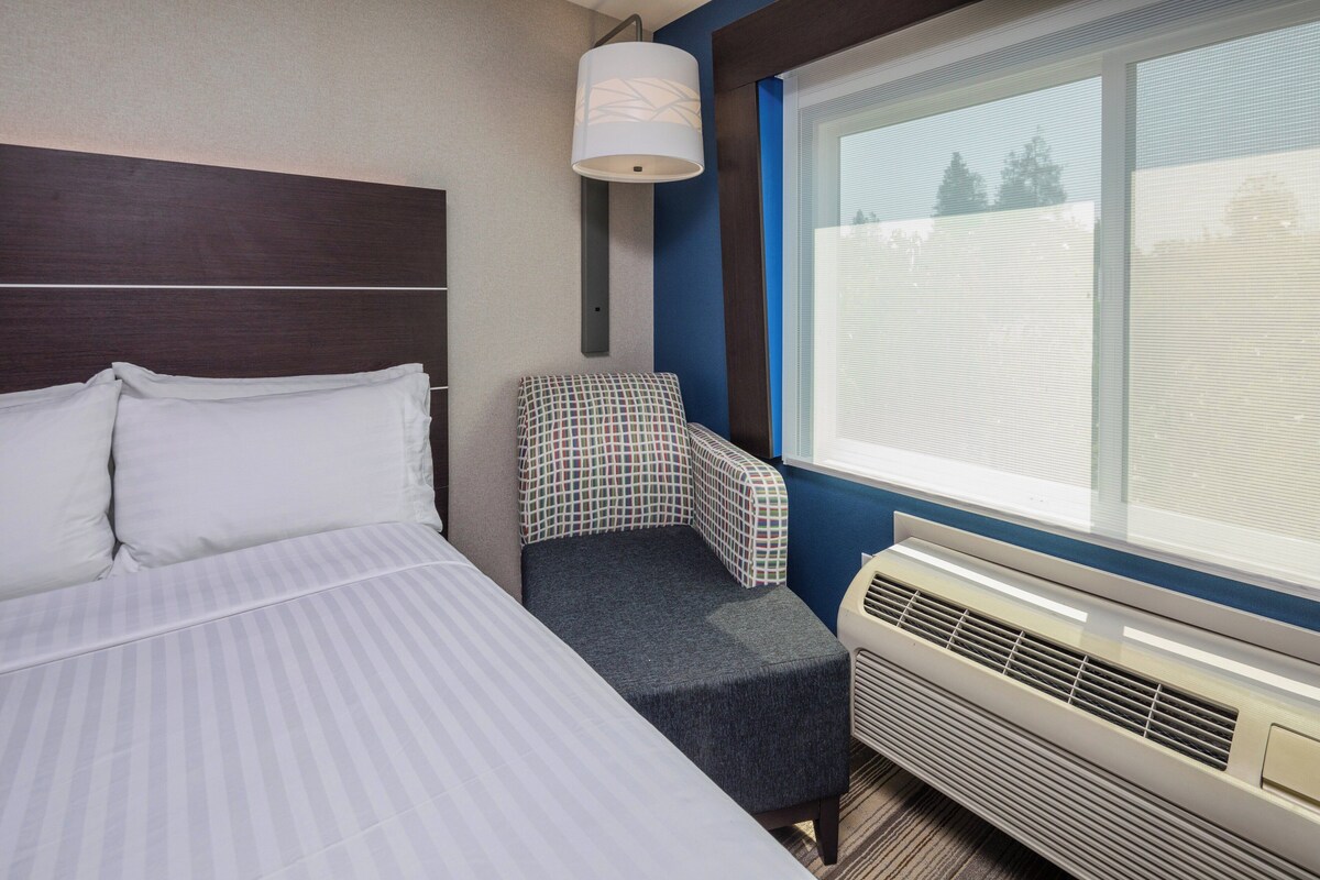 A hotel room with a bed, a chair, a window, and an air conditioning unit.