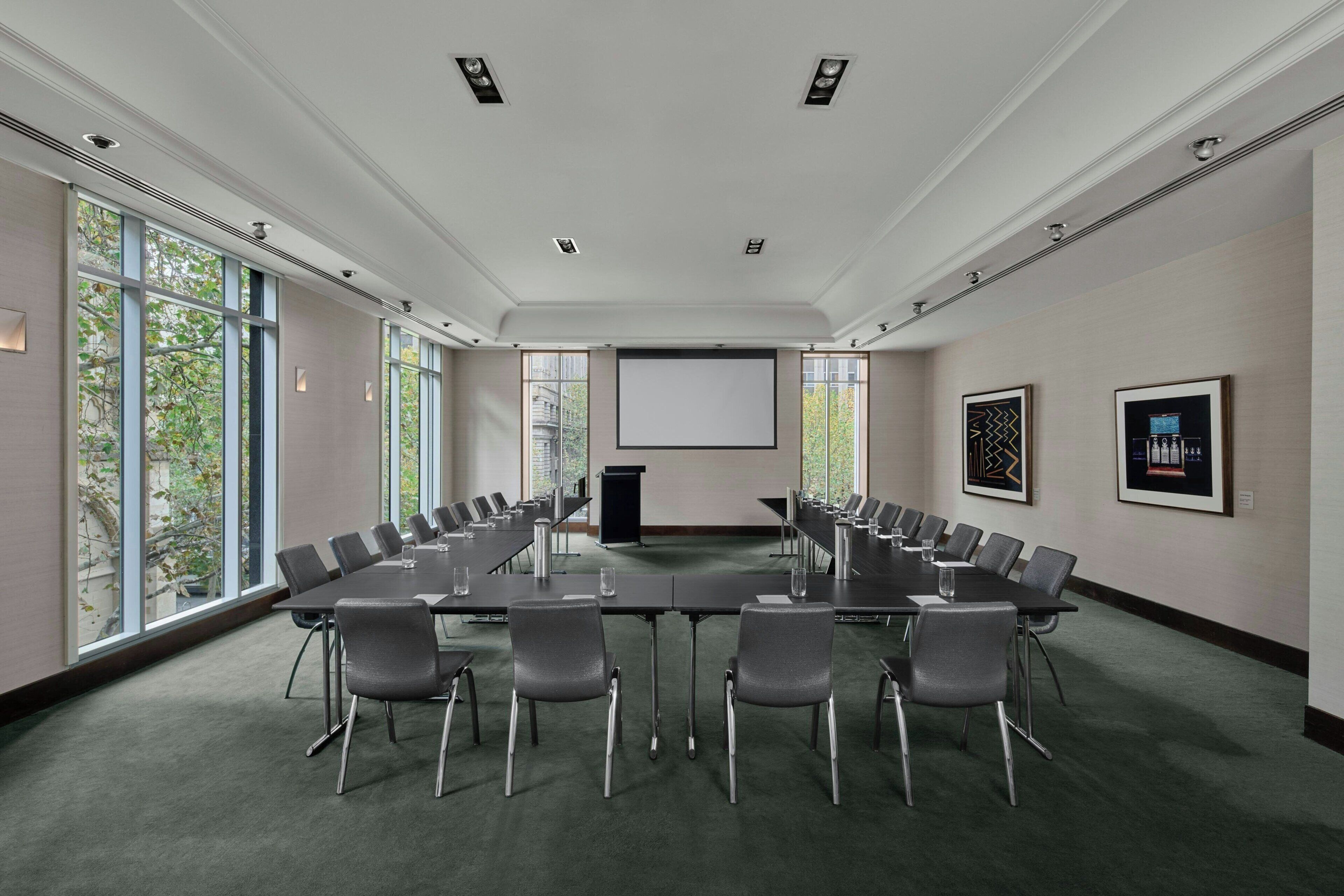 Un salón de conferencias con una larga mesa, sillas, una pantalla de proyector y ventanas amplias.