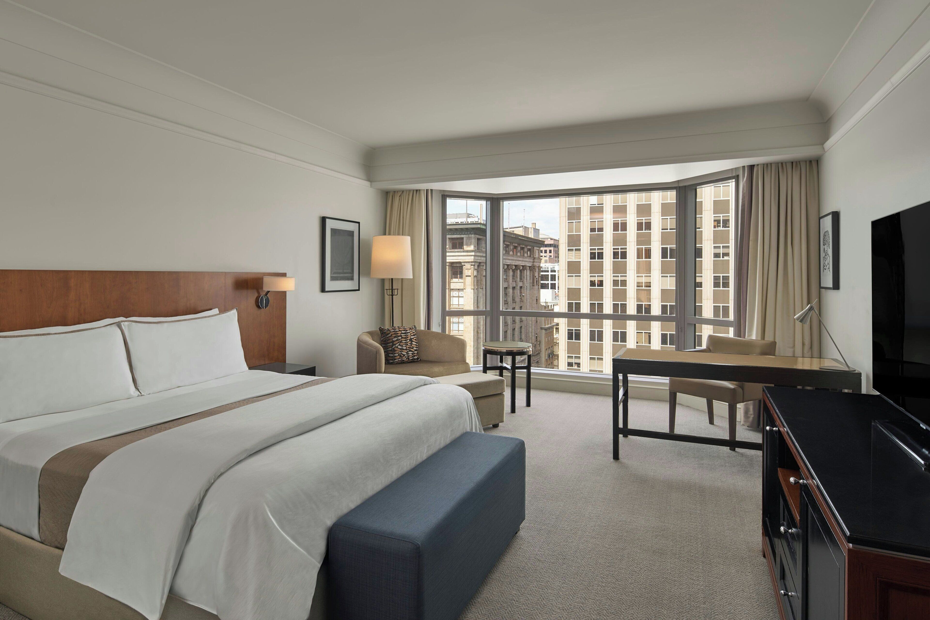 A hotel room with a large bed, a desk, a chair, a TV, and a view of the city through large windows.