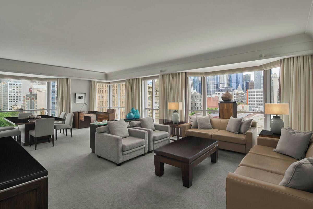 Amplia habitación de hotel con un gran ventanal que ofrece vistas a la ciudad, un comedor y una sala de estar con sofás y una mesa de centro.