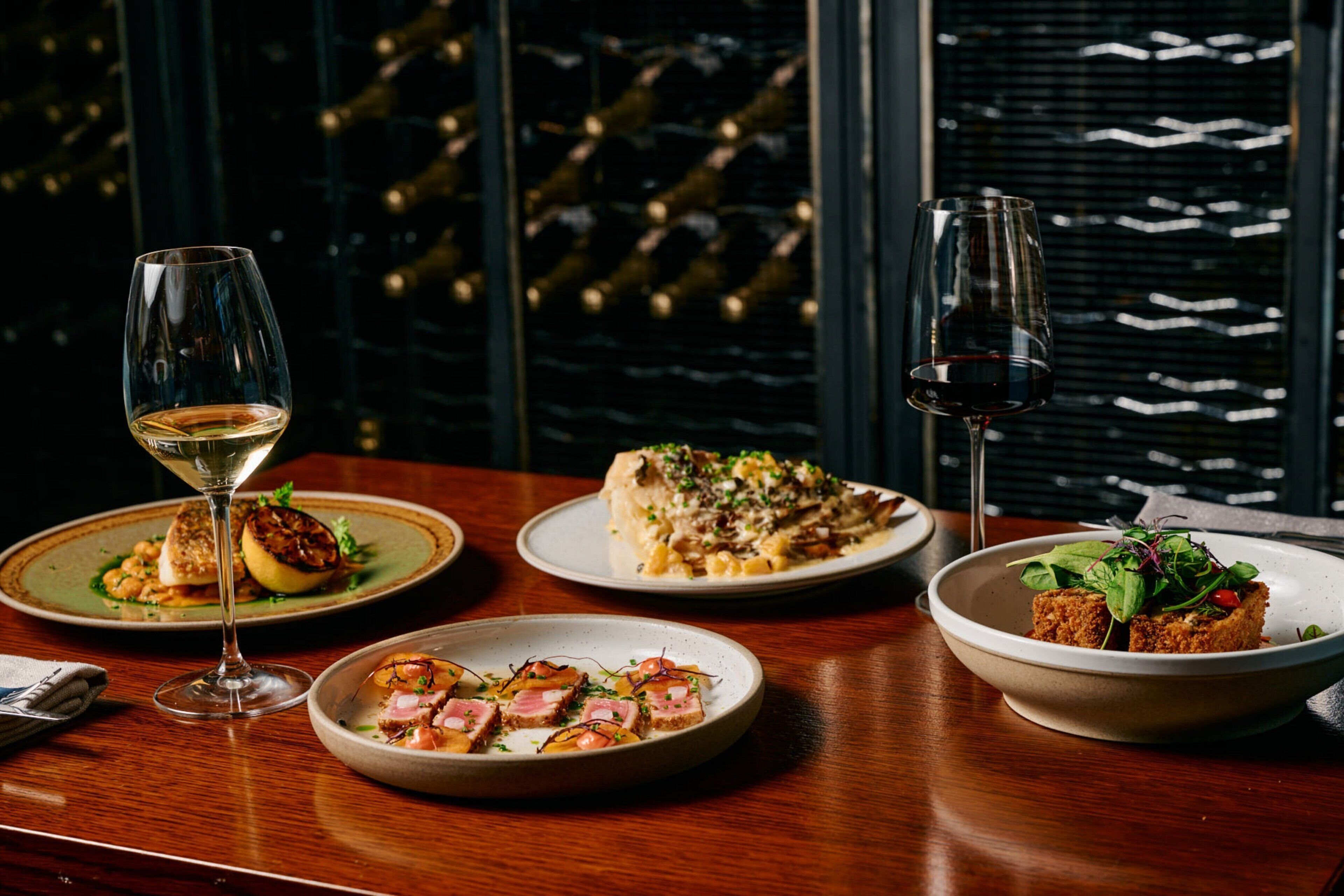 A dining table with three plates of food, two wine glasses, and a view of a wine cellar through a window.