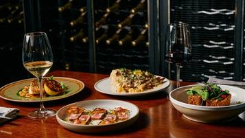 A dining table with three plates of food, two wine glasses, and a view of a wine cellar through a window.