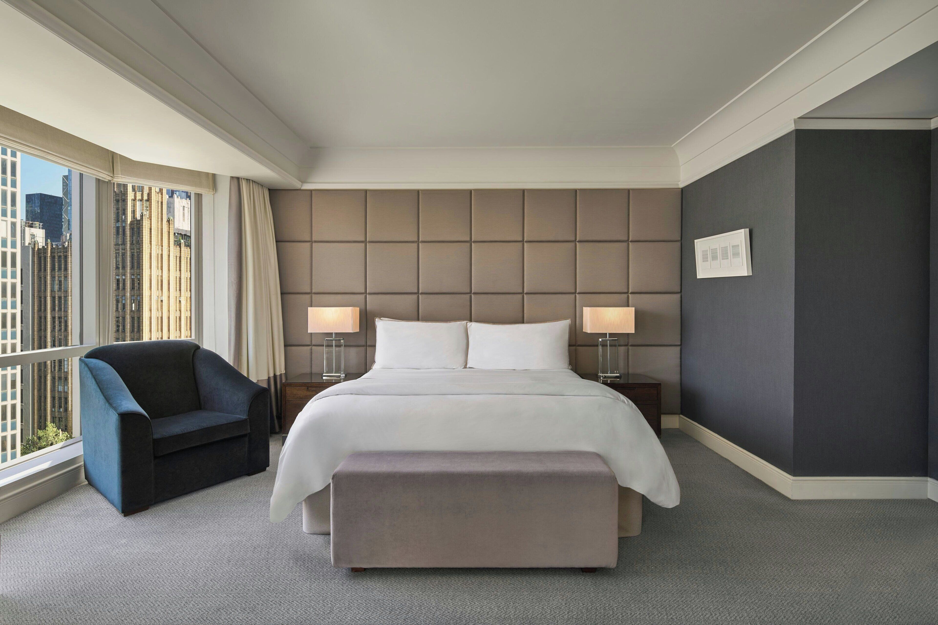 Ein Hotelzimmer mit einem großen Bett, zwei Nachttischlampen, einem Sessel und Blick auf die Stadt durch große Fenster.