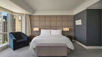 Ein Hotelzimmer mit einem großen Bett, zwei Nachttischlampen, einem Sessel und Blick auf die Stadt durch große Fenster.