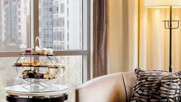 A room with a leather sofa, a three-tiered dessert stand, a floor lamp, and a view of tall buildings through large windows.