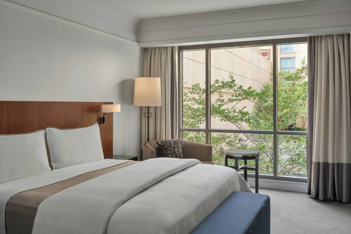 Una habitación de hotel con un ventanal grande, una cama con cabecero, una mesita de noche, una lámpara y una silla.