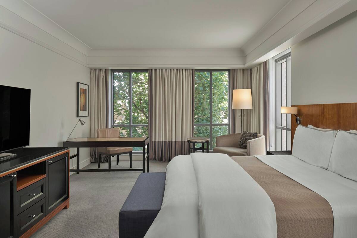 A hotel room with a large bed, a desk, a chair, a TV, and a window with curtains.