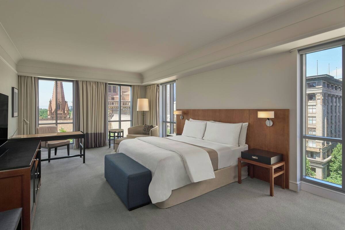 Habitación de hotel con una cama grande, un escritorio, una silla, un televisor y vista a un edificio desde la ventana.