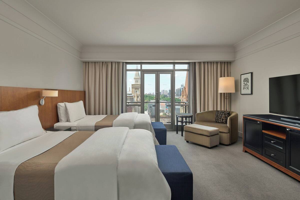 Habitación de hotel con dos camas, un sofá, un televisor y un ventanal con vistas a la ciudad.