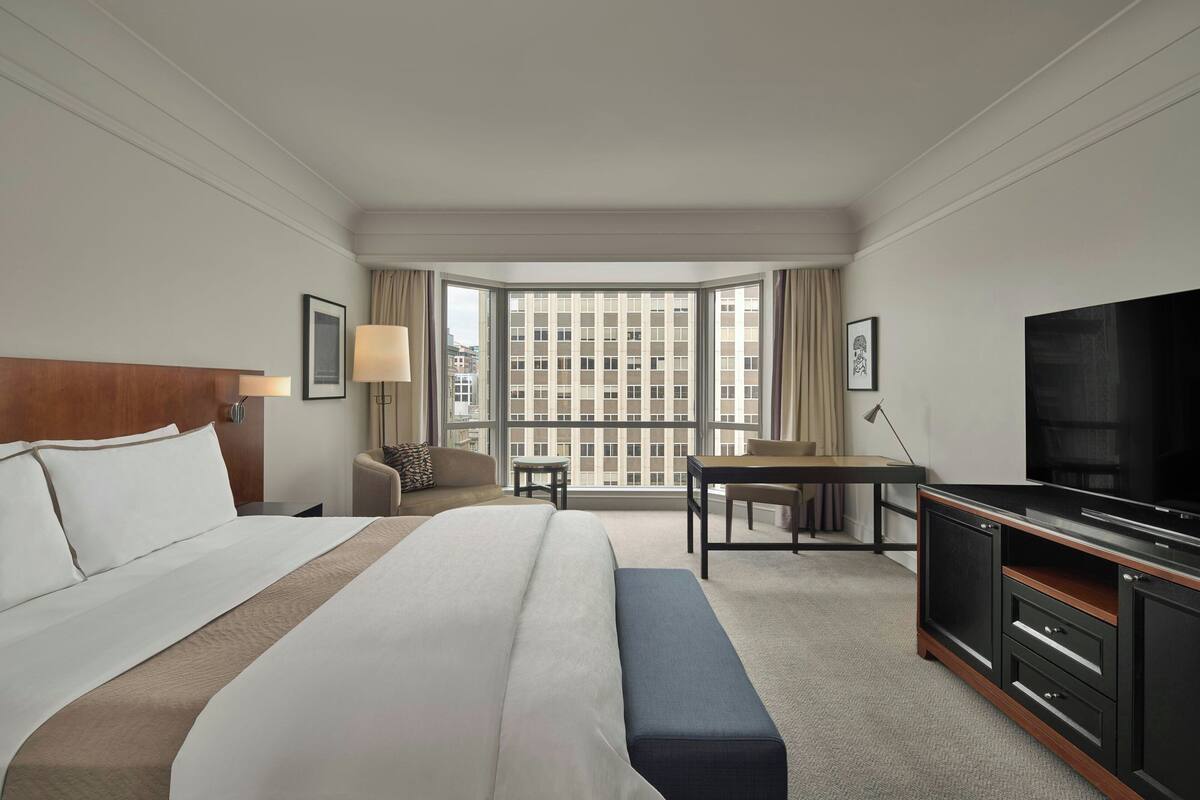 Una habitación de hotel con una cama grande, un escritorio, una televisión y vistas de la ciudad a través de amplias ventanas.