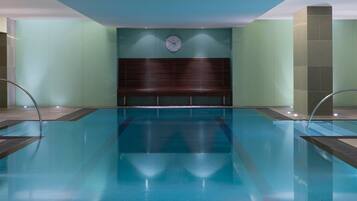 A clear indoor swimming pool with a wooden headboard, a clock on the wall, and a tiled floor.