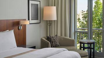 Habitación de hotel con cama, una silla, una mesita, una lámpara y vistas a zonas verdes por la ventana.