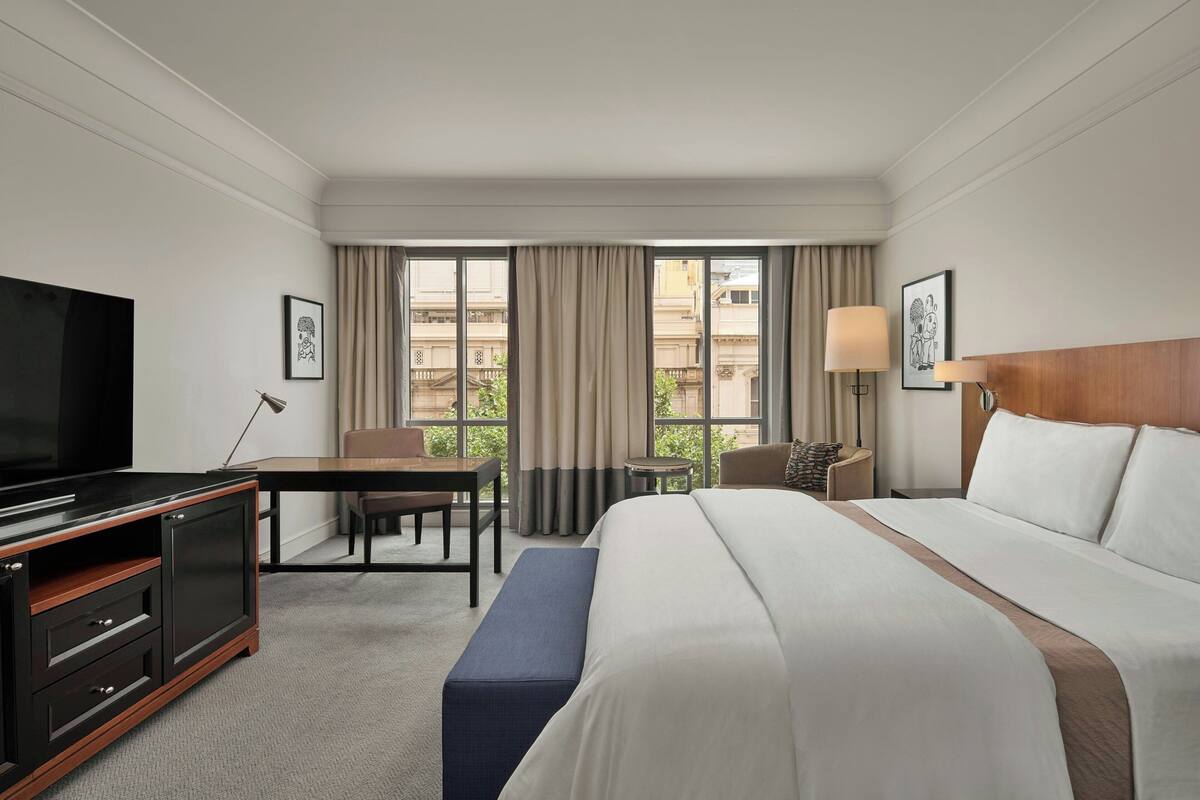 Habitación de hotel con una cama grande, un escritorio con silla, televisión y ventana con cortinas.