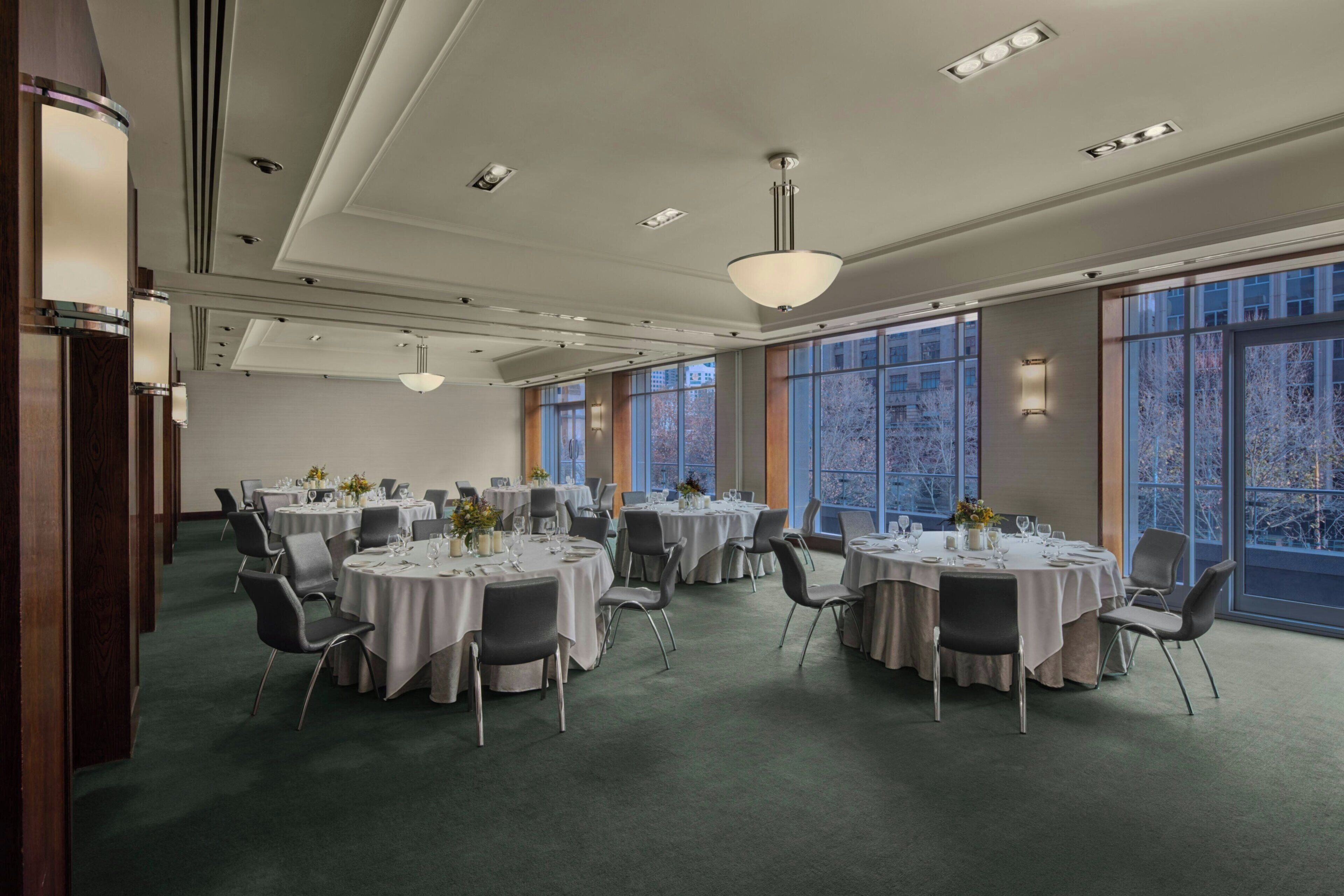 Un salón de conferencias con varias mesas redondas, cada una con sillas y cubertería, amplias ventanas que ofrecen vistas al exterior y un techo moderno con iluminación empotrada.