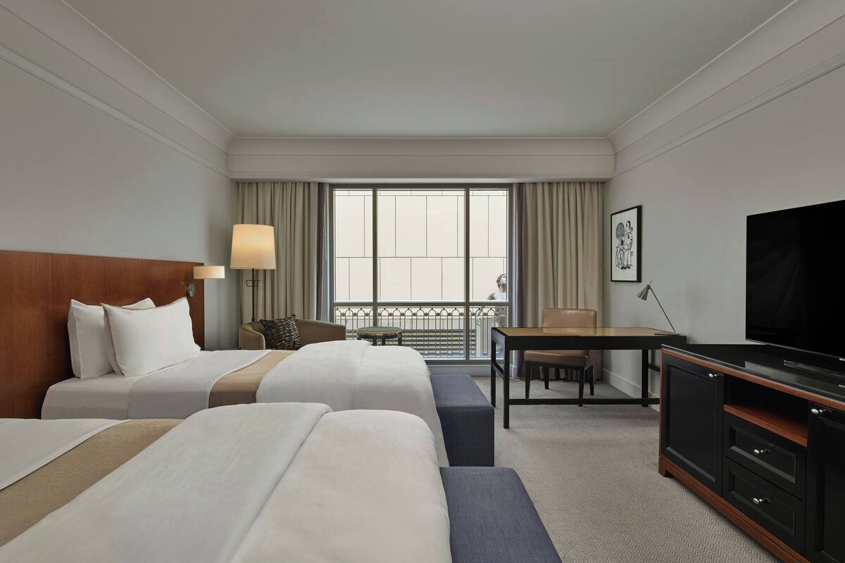 Habitación de hotel con dos camas, un escritorio, televisión y vistas al balcón.