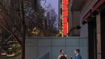 A couple dining at an outdoor table with a city street view, including a bus and a building with illuminated signage.