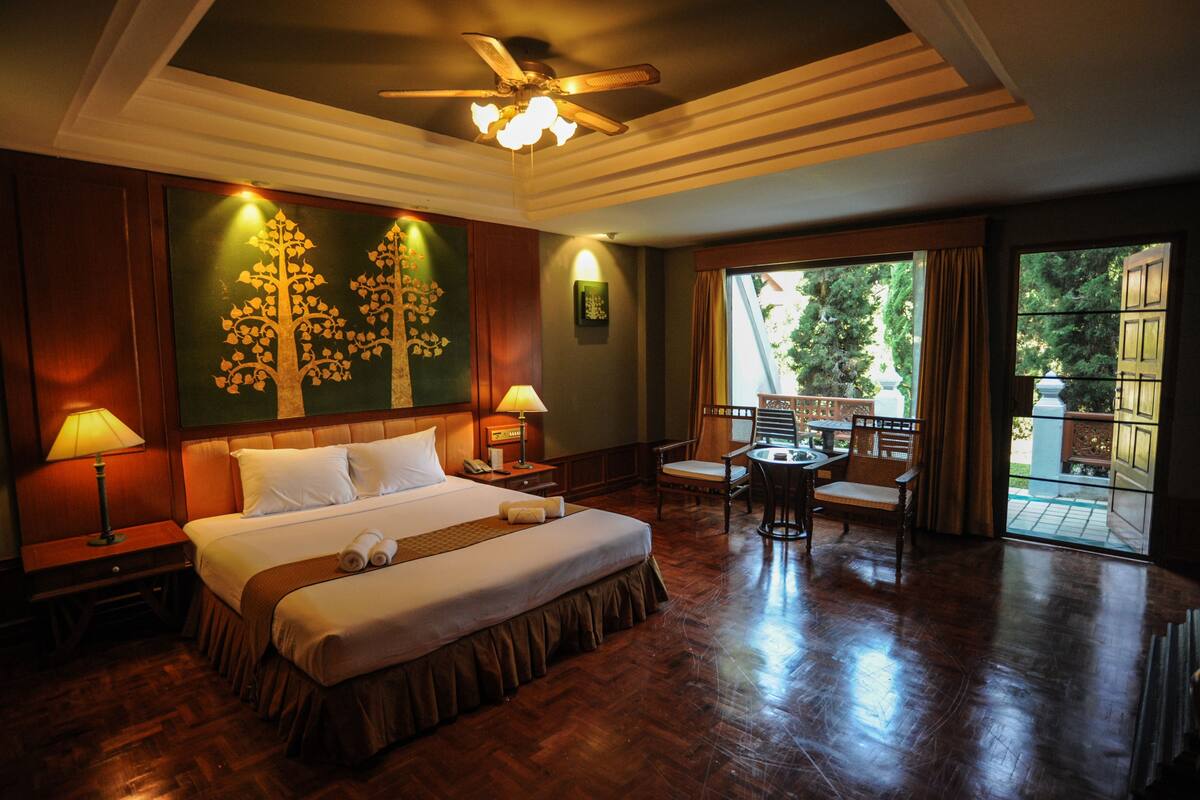 A hotel room with a large bed, two bedside lamps, a ceiling fan, a dining area with a table and chairs, and a view of the outdoors through a large window.