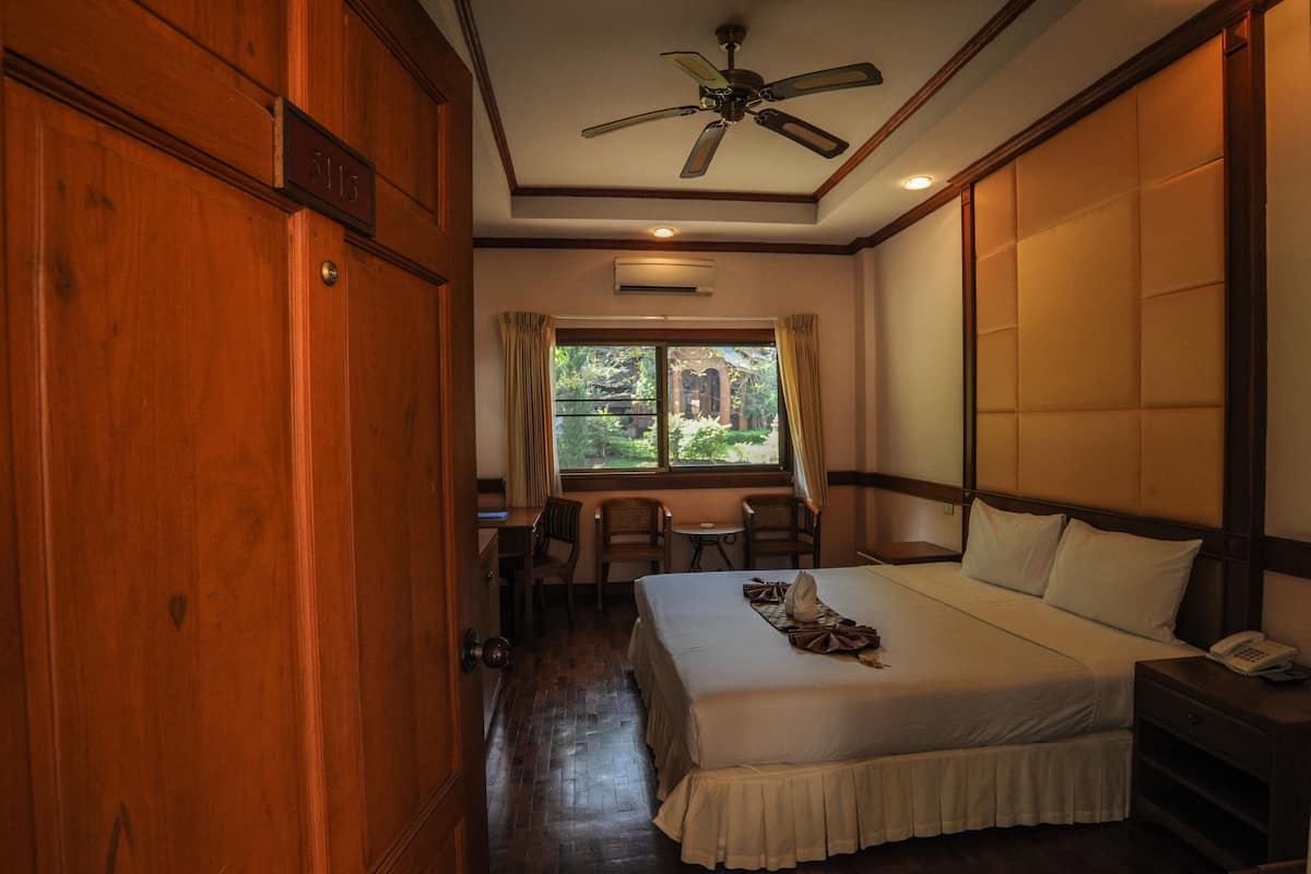 A hotel room with a large bed, a desk, two chairs, a ceiling fan, and a window with curtains.