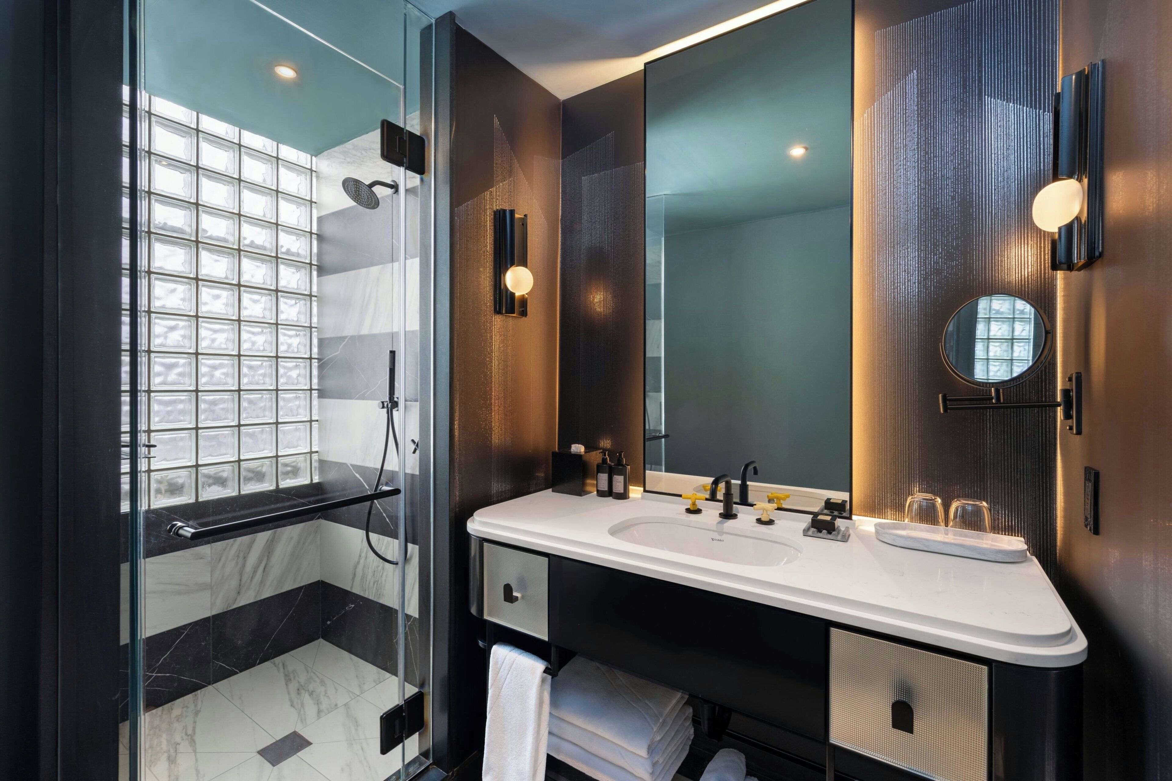 Un baño moderno con cabina de ducha de vidrio, un lavabo blanco y encimera oscura, y un espejo grande.