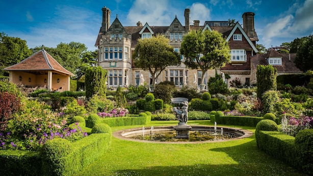 Un edificio grande e histórico con un jardín formal, que incluye una fuente y setos bien cuidados.