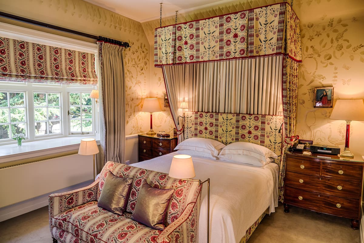 A bedroom with a four-poster bed, a nightstand with a lamp, a dresser, and a window with curtains.