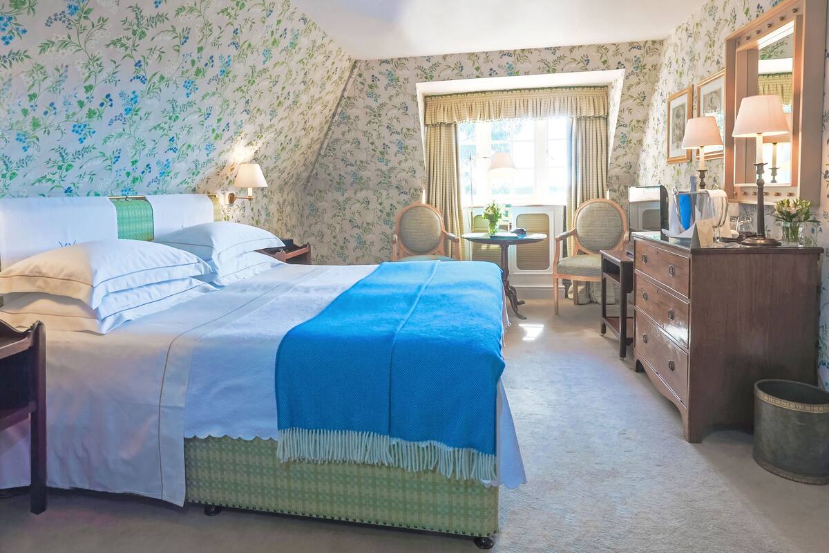 A bedroom with a bed, two chairs, a dresser, a mirror, and a window with curtains.