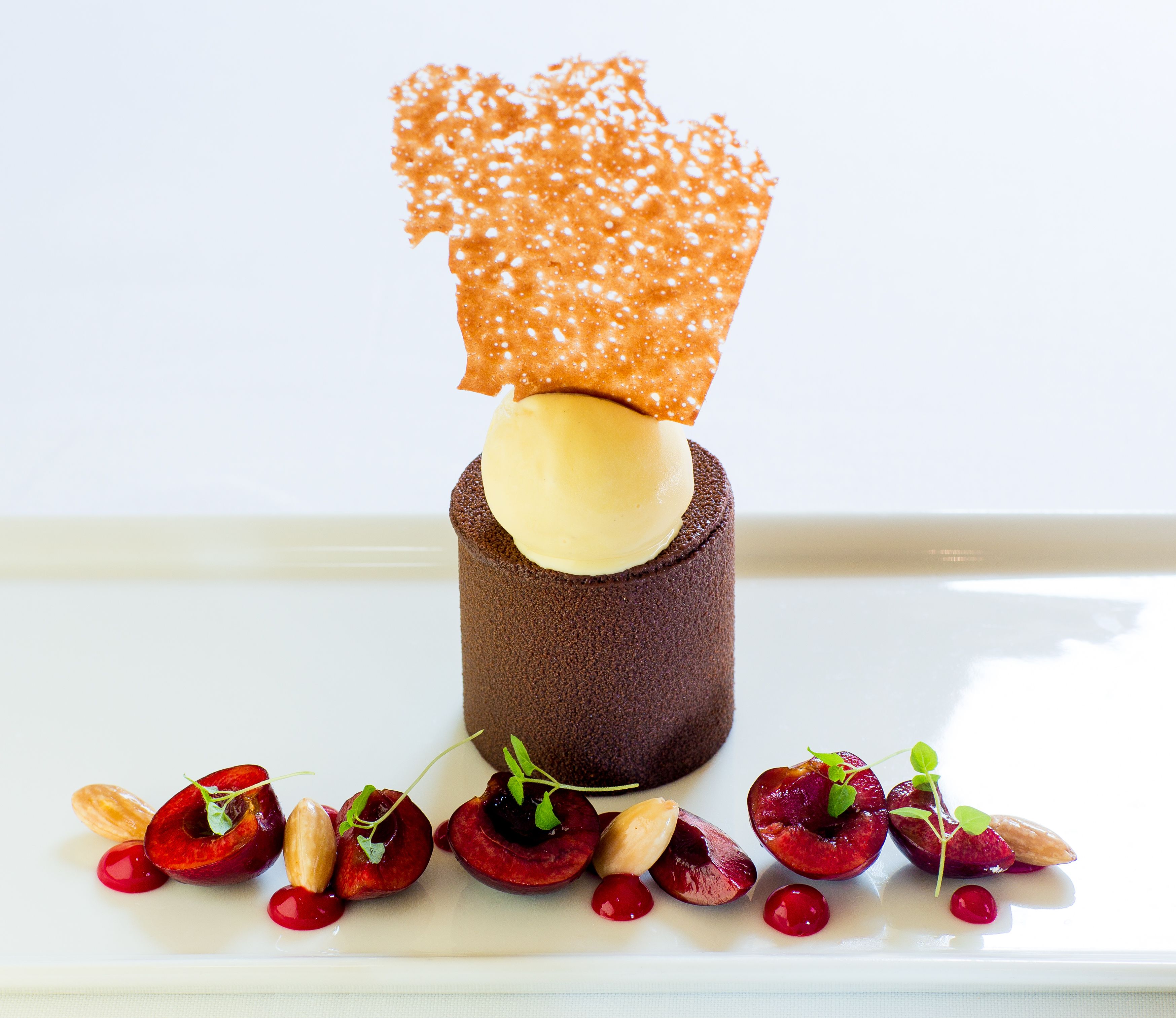 En dessert med en chokoladebund, en gul kugle, og et sprødt orange låg, omgivet af kirsebær og mandler på en hvid tallerken.