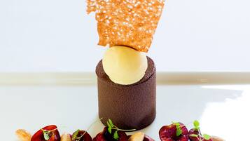 En dessert med en chokoladebund, en gul kugle, og et sprødt orange låg, omgivet af kirsebær og mandler på en hvid tallerken.