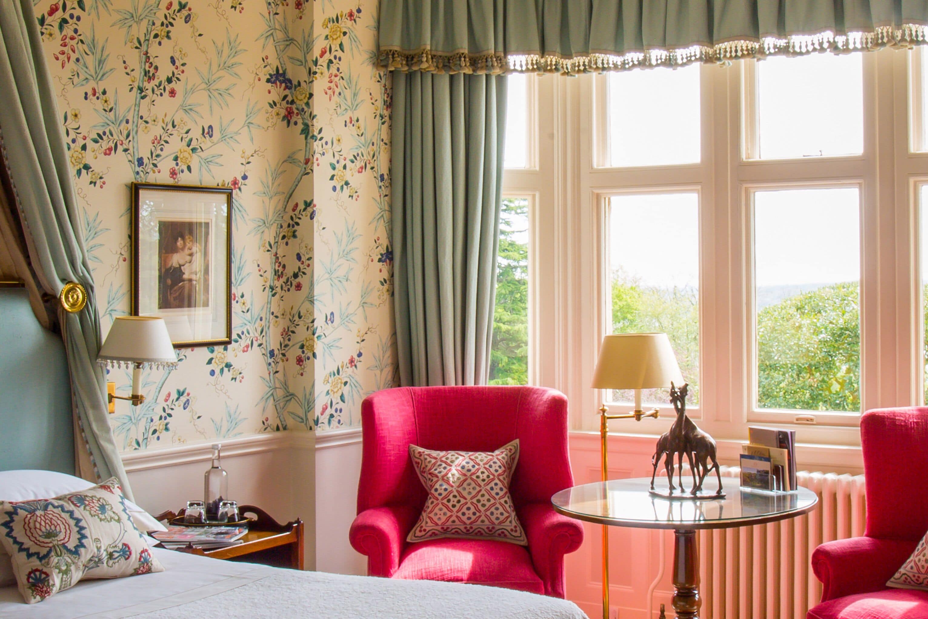 A room with floral wallpaper, a red armchair, a round table with a lamp, and a window with a view of greenery.