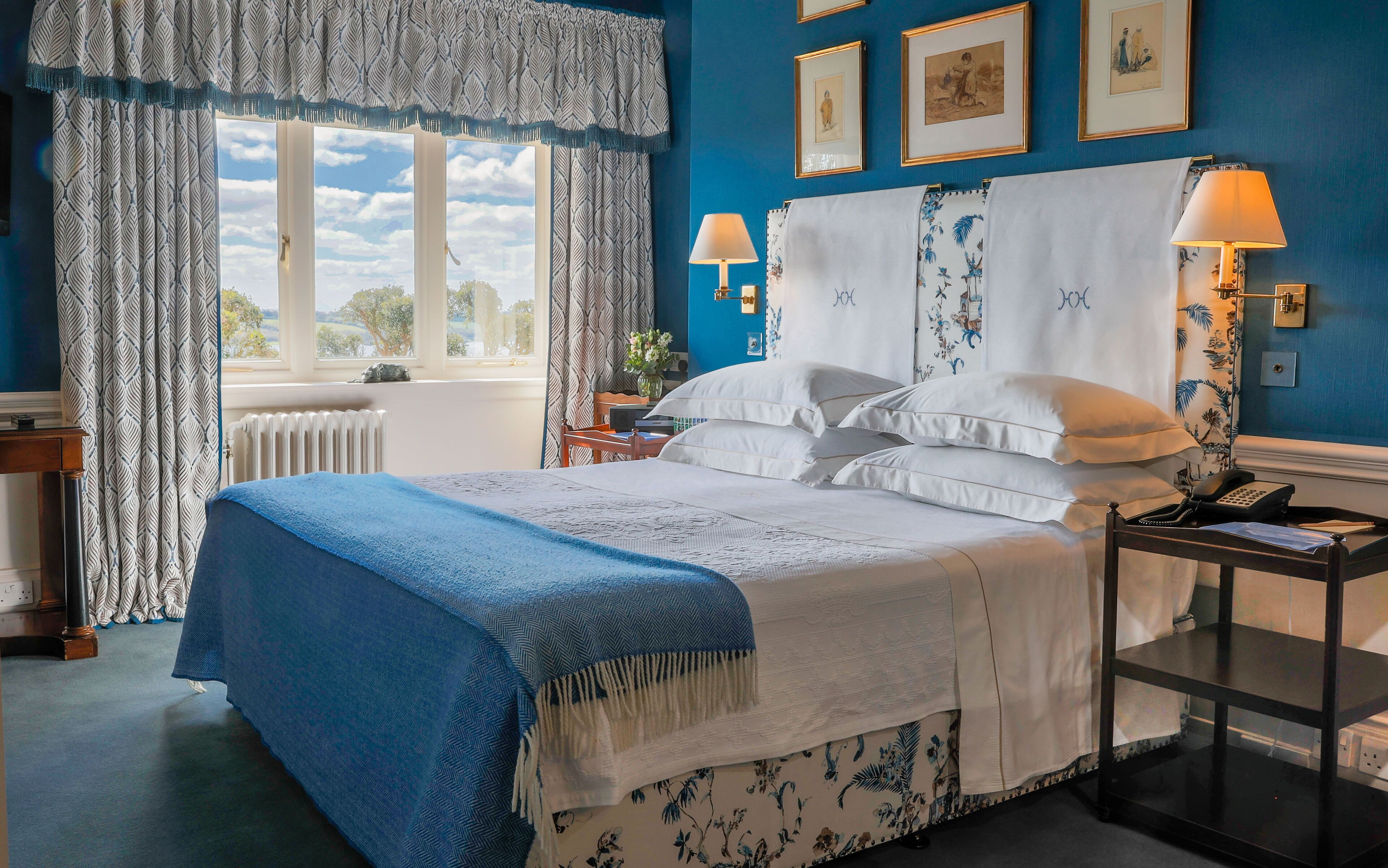A neatly arranged bedroom with a bed, two bedside lamps, a nightstand, a window with curtains, and framed pictures on the wall.