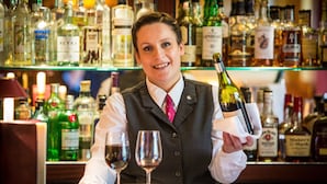 Un cantinero sostiene una botella de vino y una copa, con una barra repleta de diferentes botellas de licores al fondo.