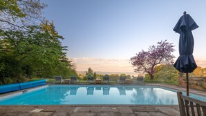 Una piscina con sillas de descanso y una sombrilla, rodeada de árboles y un cielo despejado.