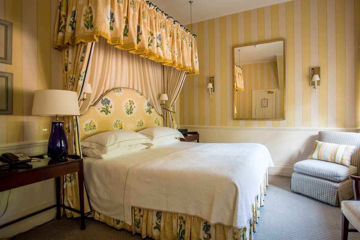 A hotel room with a bed, a chair, a desk with a lamp, and a mirror on the wall.