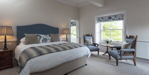 A bedroom with a large bed, two chairs, a small table, and a window with blue patterned curtains. - Farlam Hall Hotel & Restaurant (Brampton)