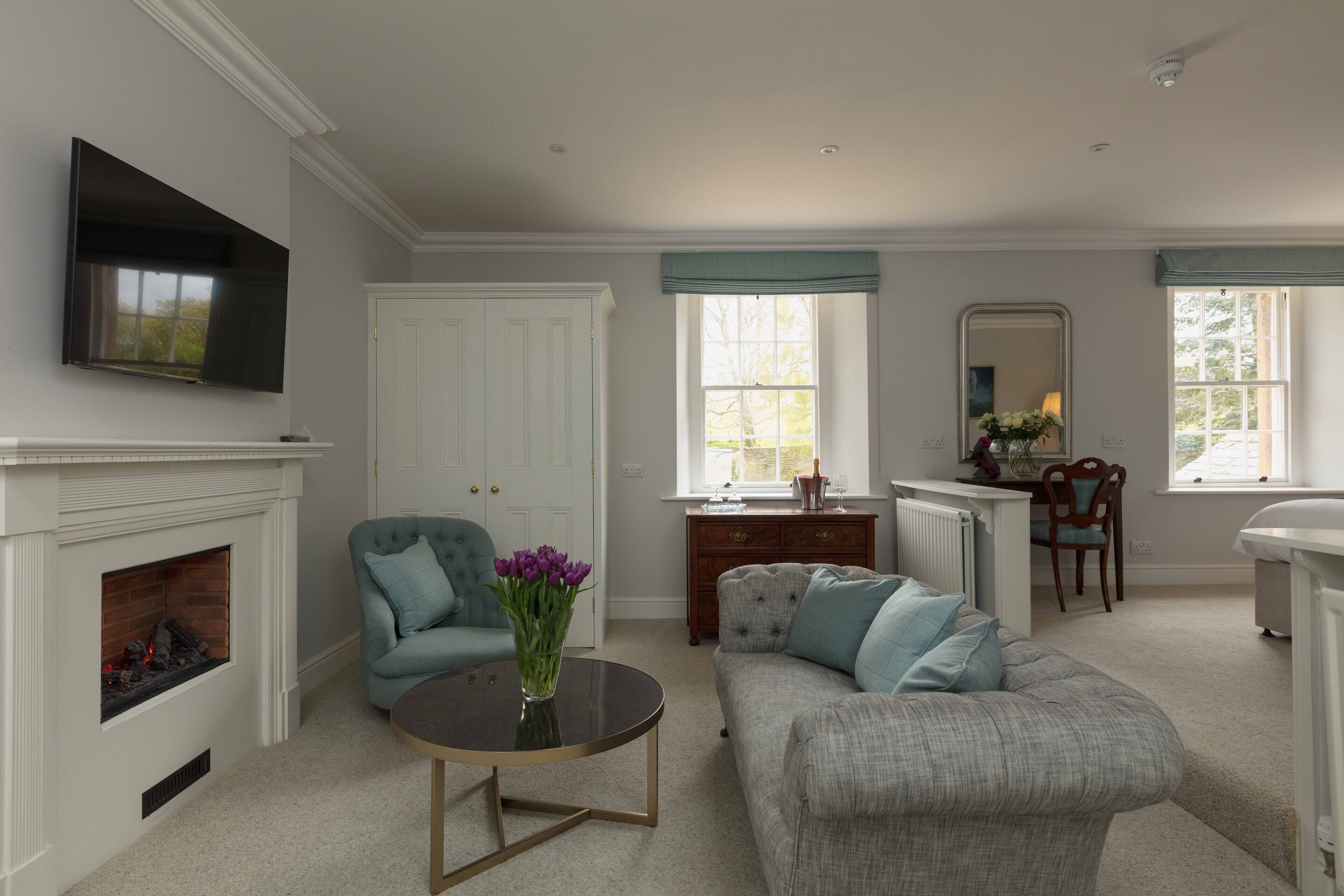 A living room with a fireplace, a television mounted on the wall, a sofa, a coffee table with a vase of flowers, and a window with a view of trees.
