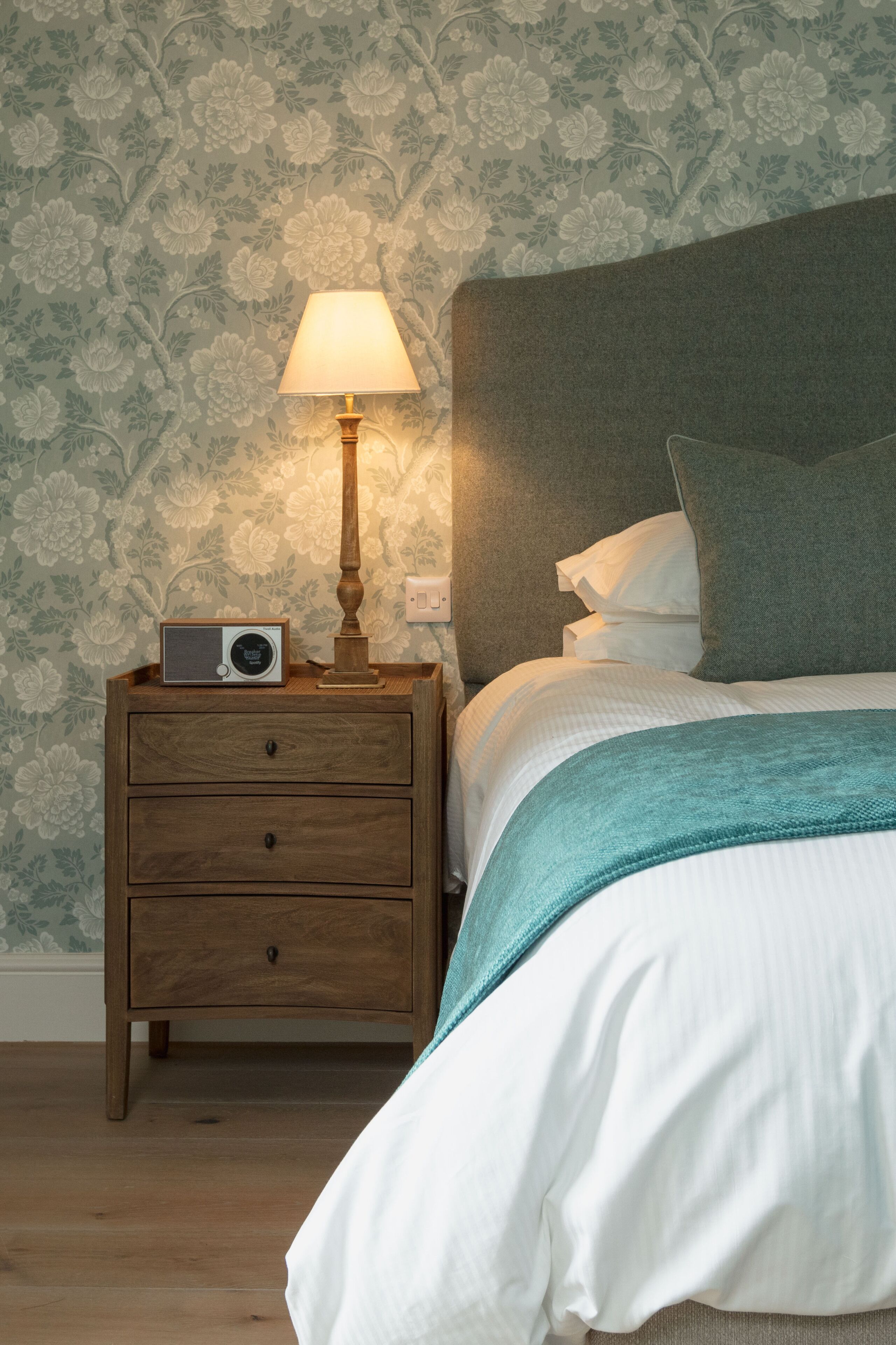 A neatly made bed with a teal throw, a wooden nightstand with a radio, and a lamp.